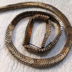 May include: A gold-toned chevron-shaped beaded necklace. The beads are arranged in a zig-zag pattern and are connected by a thin string.
