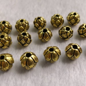 May include: A collection of fourteen small, round, gold-colored metal beads. Each bead features a textured, layered petal design resembling a flower bud or artichoke, with a central hole running through the middle for threading purposes.