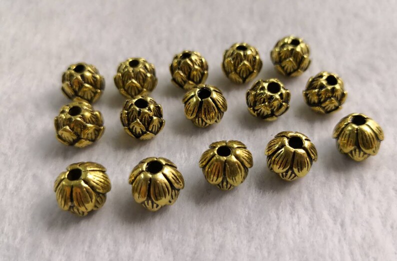 May include: A collection of fourteen small, round, gold-colored metal beads. Each bead features a textured, layered petal design resembling a flower bud or artichoke, with a central hole running through the middle for threading purposes.