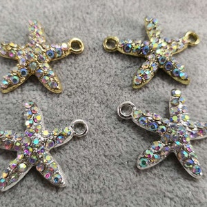 Op de afbeelding: Vier bedels in de vorm van een zeester met iriserende strass-steentjes in metalen zettingen. Twee bedels hebben goudkleurige metalen zettingen en twee hebben zilverkleurige metalen zettingen. Elke zeester heeft twee kleine ronde oogjes.