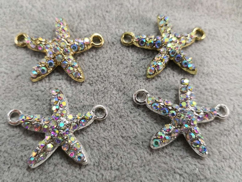 Op de afbeelding: Vier bedels in de vorm van een zeester met iriserende strass-steentjes in metalen zettingen. Twee bedels hebben goudkleurige metalen zettingen en twee hebben zilverkleurige metalen zettingen. Elke zeester heeft twee kleine ronde oogjes.