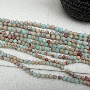May include: Strands of round gemstone beads in varying sizes. The beads are a mix of muted colors, including light blue, beige, and reddish-brown. The beads are strung together, ready for jewelry making or crafting.