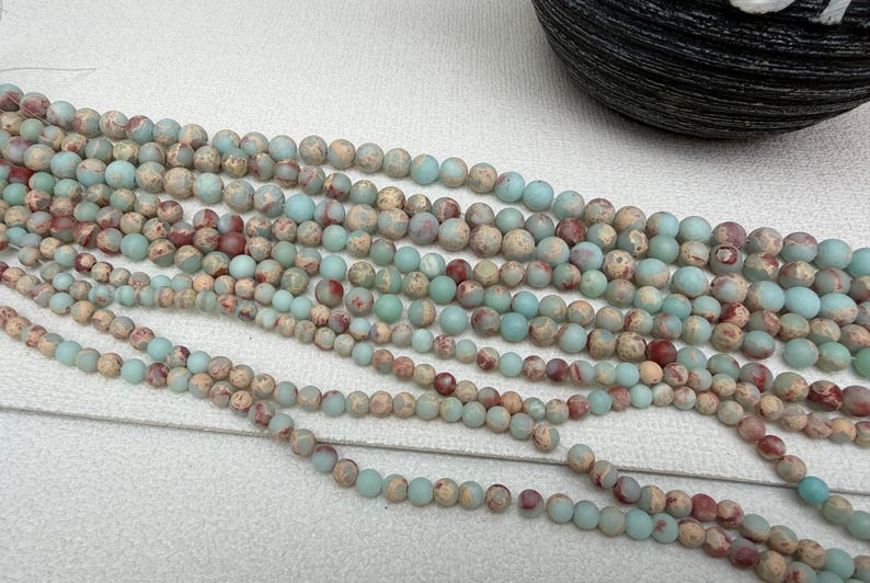 May include: Strands of round gemstone beads in varying sizes. The beads are a mix of muted colors, including light blue, beige, and reddish-brown. The beads are strung together, ready for jewelry making or crafting.