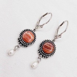 May include: A pair of silver dangle earrings with a round, orange and brown stone set in a silver bezel. Each earring has a small white pearl dangling below the stone.
