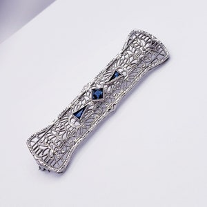May include: A silver filigree brooch with three blue sapphire stones. The brooch is in the shape of a long rectangle with a delicate, intricate design.