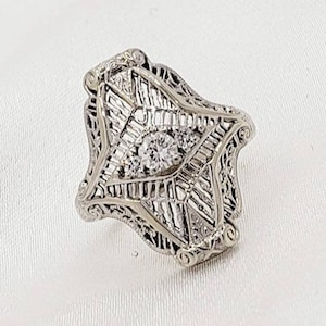 May include: A silver filigree ring with a large, clear, round-cut diamond in the center. The ring is set with smaller diamonds around the center stone.