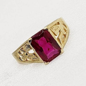 May include: A gold-toned ring featuring a large, rectangular, deep red gemstone. The ring's band has an ornate, openwork design with a detailed pattern. The gemstone is the focal point, set prominently in the center.