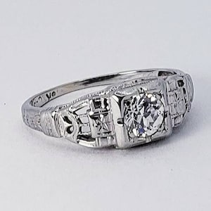 May include: A silver engagement ring with a single round diamond set in a filigree design.