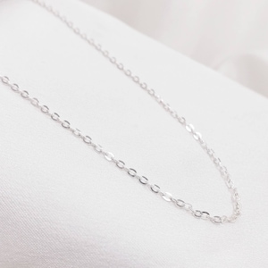May include: A delicate silver chain necklace with an oval link design. The necklace is displayed on a white, silky background, highlighting its simple elegance. The chain is a fine piece of jewelry.