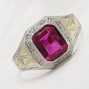 May include: A ring with a large, emerald-cut ruby set in a gold and silver band. The band is decorated with intricate floral designs.