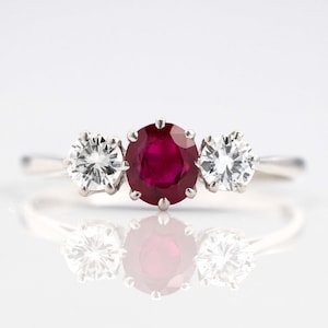 May include: A three-stone ring featuring a large, oval-cut ruby center stone flanked by two smaller, round, clear gemstones. The ring has a silver band and is set against a white background, with a reflection visible below.