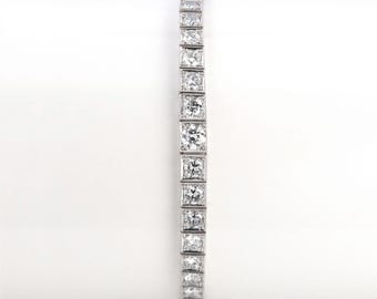 May include: A silver-toned tennis bracelet featuring a continuous row of square-set, clear, faceted stones. The design consists of individual square metal links, each holding a single brilliant-cut stone, creating a uniform and linear aesthetic.