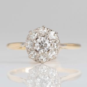 Puede incluir: Un anillo de compromiso vintage con un grupo de diamantes redondos engastados en un halo de metal blanco. El anillo tiene una banda de oro amarillo, creando un diseño de dos tonos. Los diamantes brillan sobre el fondo claro.