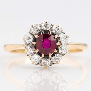 Peut inclure: Bague en or avec une pierre rouge centrale, ronde, entourée d'un halo de petites pierres claires. Le corps de la bague est en or uni. Les pierres sont serties dans un entourage détaillé et orné.