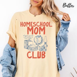 May include: Butter-colored t-shirt with the words "Homeschool Mom Club" in red. The shirt features a graphic of books, a globe, a lamp, and blocks in blue. The phrase "shaping minds with patience and coffee" is also included.
