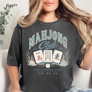 May include: A charcoal gray t-shirt featuring a graphic with the text "MAHJONG CLUB EST. 1920" in light blue and cream. The design includes three mahjong tiles, seashells, seaweed, and the text "PLAY CONNECT ENJOY" with Chinese characters below.