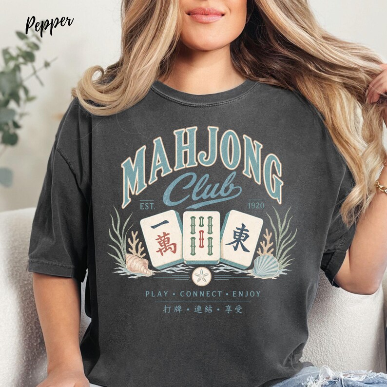 May include: A charcoal gray t-shirt featuring a graphic with the text "MAHJONG CLUB EST. 1920" in light blue and cream. The design includes three mahjong tiles, seashells, seaweed, and the text "PLAY CONNECT ENJOY" with Chinese characters below.