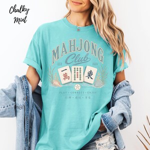 May include: A chalky mint t-shirt featuring a graphic that reads Mahjong Club Est 1920. The design includes three mahjong tiles, seashells, and the text Play Connect Enjoy with corresponding Chinese characters. The shirt has a crew neckline.