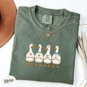 May include: A moss green crew neck t-shirt featuring a graphic of four white geese standing in a row. Each goose holds a different mahjong tile. The shirt has a Comfort Colors brand label at the inner neckline and a thin gold-toned chain necklace.