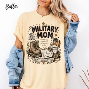 May include: A butter yellow t-shirt featuring a graphic with the text MILITARY MOM, always in my heart, always my hero, PRAYING, SUPPORTING, LOVING. The design includes dog tags, combat boots, an American flag, a mug labeled proud mama, and letters.