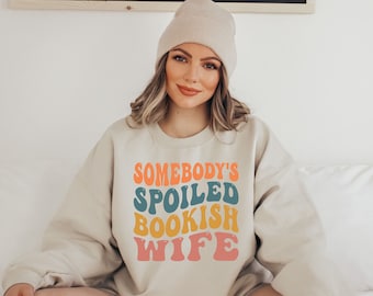 May include: A beige sweatshirt with the text "Somebody's Spoiled Bookish Wife" in colorful retro-style lettering.