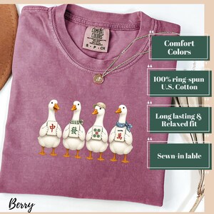 May include: A berry-colored crew neck t-shirt featuring a graphic of four white ducks, each holding a different Mahjong tile. The shirt has a Comfort Colors brand label at the inner neckline and a thin silver-toned chain necklace resting on the fabric.