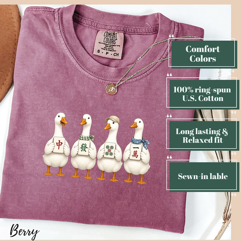 May include: A berry-colored crew neck t-shirt featuring a graphic of four white ducks, each holding a different Mahjong tile. The shirt has a Comfort Colors brand label at the inner neckline and a thin silver-toned chain necklace resting on the fabric.