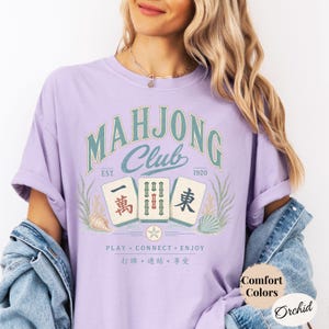 May include: An orchid-colored t-shirt featuring a graphic with the text Mahjong Club Est. 1920, Play Connect Enjoy, and Chinese characters. The design includes three mahjong tiles, seaweed, and seashells. A circular label reads Comfort Colors Orchid.