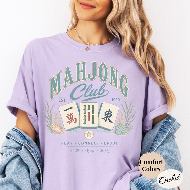 May include: An orchid-colored t-shirt featuring a graphic with the text Mahjong Club Est. 1920, Play Connect Enjoy, and Chinese characters. The design includes three mahjong tiles, seaweed, and seashells. A circular label reads Comfort Colors Orchid.