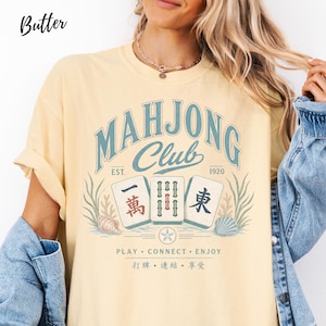 May include: A butter yellow short-sleeve t-shirt featuring a graphic with the text "MAHJONG CLUB EST. 1920" in blue. The design includes three mahjong tiles, seashells, and seaweed, with the text "PLAY &bull; CONNECT &bull; ENJOY" and Chinese characters below.