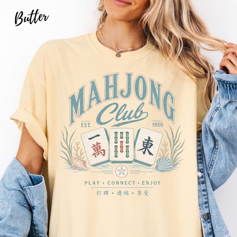 May include: A butter yellow short-sleeve t-shirt featuring a graphic with the text "MAHJONG CLUB EST. 1920" in blue. The design includes three mahjong tiles, seashells, and seaweed, with the text "PLAY &bull; CONNECT &bull; ENJOY" and Chinese characters below.