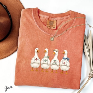 May include: A terracotta-colored crew neck t-shirt featuring a graphic of four white ducks standing in a row. Each duck holds a mahjong tile with Chinese characters or green dots. The shirt has a Comfort Colors label at the inner neckline.