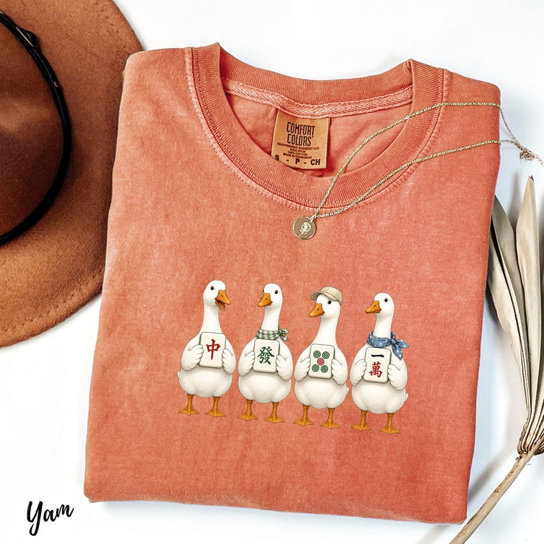 May include: A terracotta-colored crew neck t-shirt featuring a graphic of four white ducks standing in a row. Each duck holds a mahjong tile with Chinese characters or green dots. The shirt has a Comfort Colors label at the inner neckline.