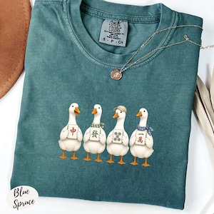 May include: A teal crewneck t-shirt featuring a graphic of four white ducks standing in a row, each holding a different mahjong tile. The shirt has a Comfort Colors label at the neck and is paired with a thin gold chain necklace.