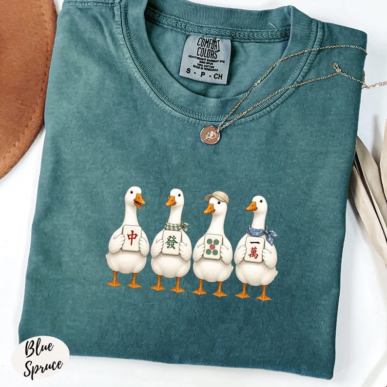 May include: A teal crewneck t-shirt featuring a graphic of four white ducks standing in a row, each holding a different mahjong tile. The shirt has a Comfort Colors label at the neck and is paired with a thin gold chain necklace.