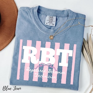 May include: A blue jean colored t-shirt featuring a graphic with vertical light pink stripes and the white text RBT. Below the letters is a pink bow graphic and the white script text Registered Behavior Technician. The shirt has a Comfort Colors label.