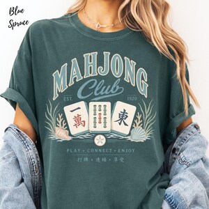 May include: A blue spruce t-shirt featuring a graphic with the text "MAHJONG Club EST. 1920" above three mahjong tiles, seashells, and seaweed. Below the tiles, the text reads "PLAY CONNECT ENJOY" followed by Chinese characters.