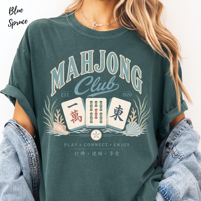 May include: A blue spruce t-shirt featuring a graphic with the text "MAHJONG Club EST. 1920" above three mahjong tiles, seashells, and seaweed. Below the tiles, the text reads "PLAY CONNECT ENJOY" followed by Chinese characters.