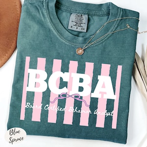 Peut inclure: Un t-shirt Comfort Colors bleu sarcelle avec le texte "BCBA" en lettres blanches, des rayures verticales roses et les mots "Board Certified Behavior Analyst". Un collier en or avec un pendentif est posé sur le t-shirt.