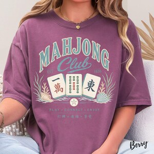May include: A berry-colored t-shirt featuring the text "MAHJONG Club" in light blue, "EST 1920", and three mahjong tiles. The design includes illustrations of seashells and seaweed, with the text "PLAY CONNECT ENJOY" and Chinese characters below.