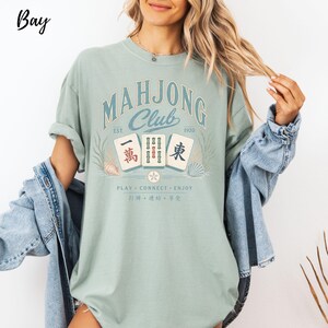 May include: A sage green t-shirt featuring a graphic with the text "MAHJONG CLUB EST. 1920" and "PLAY CONNECT ENJOY". The design includes three mahjong tiles with Chinese characters, surrounded by illustrations of seashells and seaweed in muted tones.