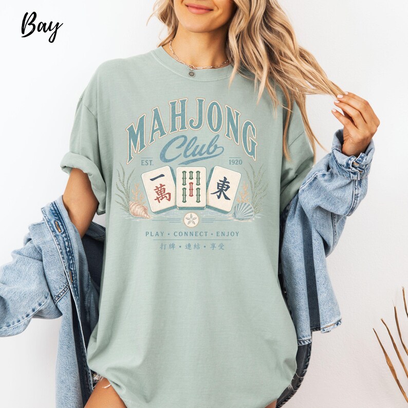May include: A sage green t-shirt featuring a graphic with the text "MAHJONG CLUB EST. 1920" and "PLAY CONNECT ENJOY". The design includes three mahjong tiles with Chinese characters, surrounded by illustrations of seashells and seaweed in muted tones.