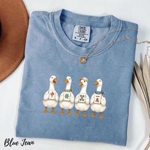 May include: A blue jean colored t-shirt featuring a graphic of four white ducks standing in a row. Each duck holds a different mahjong tile. The shirt has a crew neckline and a Comfort Colors brand label on the inside of the collar.