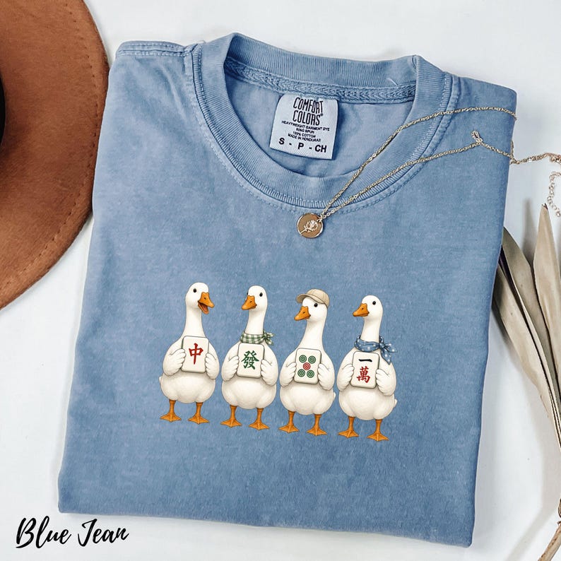 May include: A blue jean colored t-shirt featuring a graphic of four white ducks standing in a row. Each duck holds a different mahjong tile. The shirt has a crew neckline and a Comfort Colors brand label on the inside of the collar.