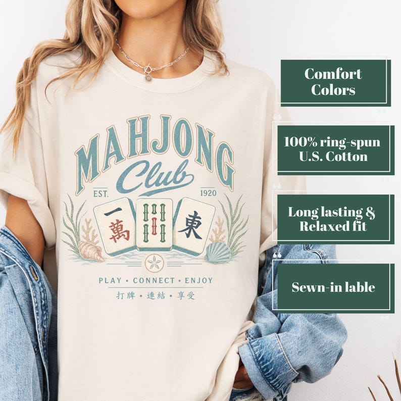 May include: A cream-colored t-shirt featuring a graphic with the text "MAHJONG Club EST. 1920" in blue, three mahjong tiles, seashells, and the text "PLAY CONNECT ENJOY" with Chinese characters. The shirt is layered under a blue denim jacket.