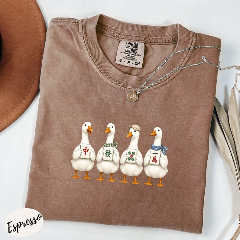 May include: A brown crew neck t-shirt featuring a graphic of four white ducks standing in a row, each holding a different Mahjong tile. The shirt has a Comfort Colors brand label inside the collar and a silver-toned necklace resting on the fabric.