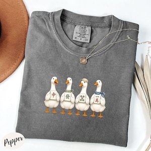 May include: A charcoal gray crew neck t-shirt featuring a graphic of four white geese standing in a row, each holding a mahjong tile. The shirt has a Comfort Colors brand label at the inner neckline and a thin gold-toned chain necklace resting on it.