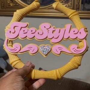 Op de afbeelding: Een grote ronde oorring met een geel frame met bamboetextuur. Het midden bevat de roze tekst TeeStyles in een cursief lettertype, geaccentueerd door roze decoratieve krullen en een klein, centraal hartje ingelegd met heldere, fonkelende steentjes.