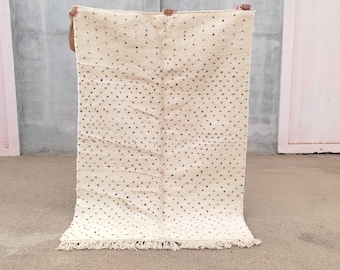 May include: A white rug with a black polka dot pattern. The rug has a fringe border and is being held up by a person's hands.