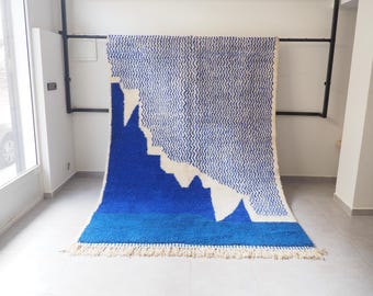 Puede incluir: Una alfombra rectangular grande con un diseño azul y blanco vibrante. La alfombra presenta un patrón similar a una montaña en tonos de azul y blanco, con flecos a lo largo del borde inferior. La alfombra se exhibe contra una pared blanca.